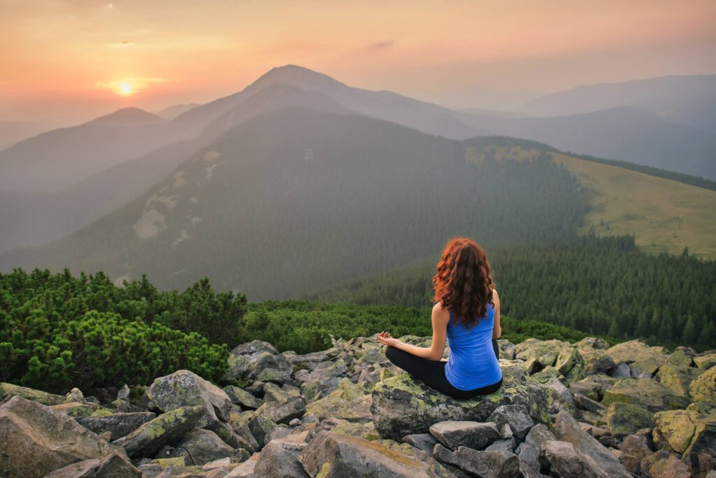 Woman with curly hair sitting on a rock overlooking a mountain with a sunrise in the back ground