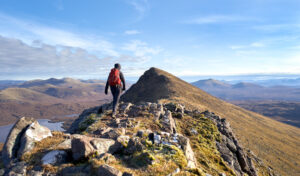 A hiker walking towards the summit of Sgurr an Tuill Bhain along a narrow rocky ridge in the Scottish Highlands on a sunny winters day.